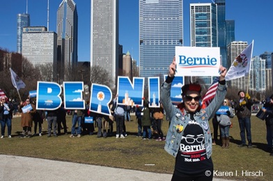 Woman holds up Bernie sign in front of a line of people spelling out large letters of Bernie